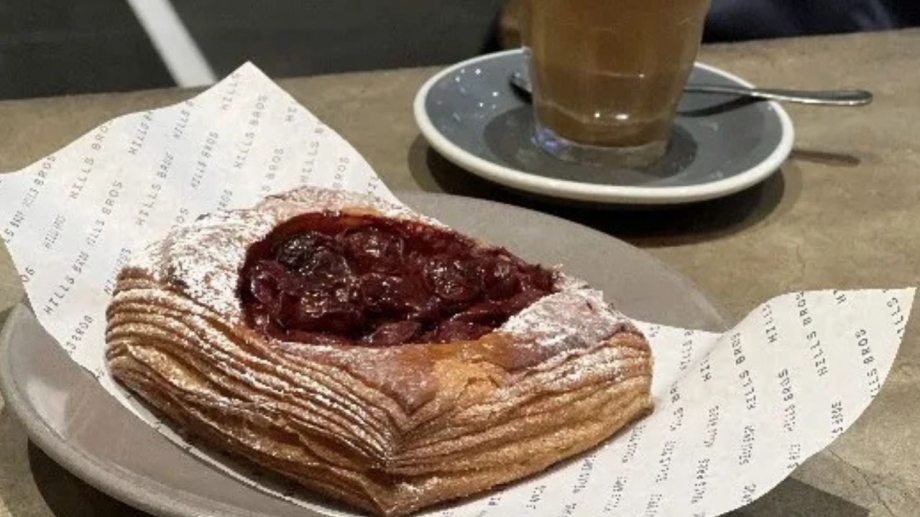 Fresh cherry Danish pastry dusted with powdered sugar, paired with a creamy latte on a saucer at Hill Bros Cafe.